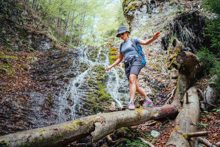 Active woman with backpack in hat dressed in active trekking clothes crossing mountain river creek with waterfall by fallen tree trunk bridge. Traveling, trekking, nature concept imageの写真素材