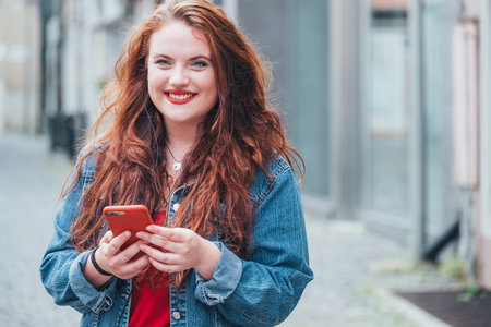 Portrait of a smiling, red-curled, long-haired Caucasian teenage girl walking down the street and video chatting on a modern smartphone. Modern people with technology devices concept image.の写真素材