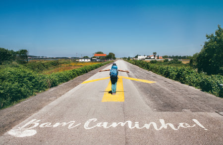 Joyful female pilgrim with backpack walking on yellow arrow road towards WAY under clear blue sky. Inspiring solo journey along famous Camino de Santiago with "Bom Caminho!" written on pavement.の写真素材