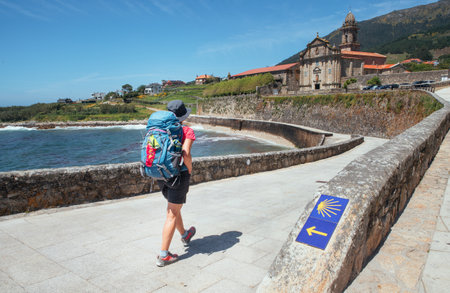 Female pilgrim with backpack walking next yellow arrow and shell directing Camino WAY inspiring solo walk on famous Camino de Santiago, Praia de Oia embankment, Royal Monastery of Santa MarÃ­a de Oiaの写真素材