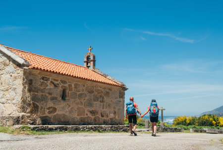 Couple of pilgrims with backpacks walking hand in hand next to old church on Camino Portuguese Way, inspiring walk on famous Camino de Santiago. Travel, pilgrimage, lifestyle, architectureの写真素材