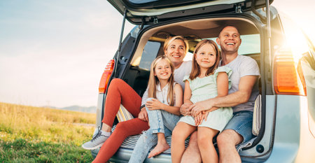 Portrait of Happy young couple with little daughters sitting inside car trunk during auto trop. They are smiling, laughing and chatting. Family values, traveling concept.の写真素材