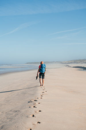 Lonely barefoot backpacker man walking by sandy Portugese beach in morning on the Atlantic ocean coast. Shot during the Camino Portuguese - popular pilgrimage route. Active people, traveling concept.の写真素材