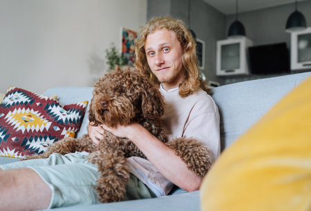 Portrait of young teenager boy with long curly hear smiling cheerfully sitting on comfortable living...