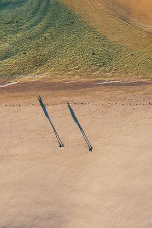 Top-down view of couple walking along sandy beach washed by Atlantic Ocean waves in Portugal casting long shadows in low sunrise light. Shot during the Camino Portuguese â popular pilgrimage routeの写真素材