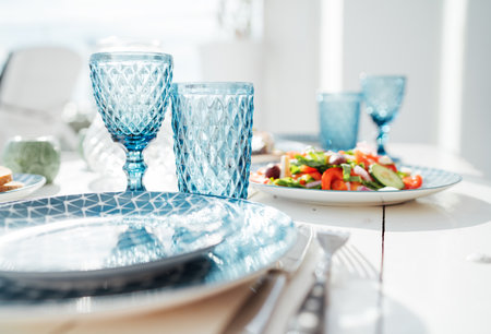 Elegant summer table setting with patterned blue glassware, ceramic plates, fresh vegetable salad,...