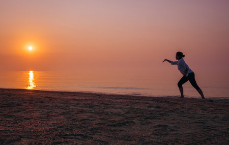 Woman practicing Tai Chi on beach at sunrise, graceful silhouette against calm sea and orange-pink sky. Mindfulness, wellness lifestyle, energy balance, harmony with nature, and body awareness conceptの写真素材