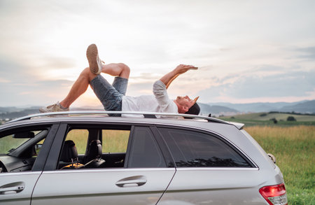 Young man lying on car roof reading a paper book at sunset. Vehicle parked on meadow overlooking scenic valley. Peaceful outdoor moment, reading hobby, education, and travel lifestyle concept.の写真素材