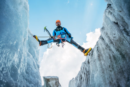 Climber in vibrant gear overcomes over crevasse on icy glacier during Lenin peak ascent under clear sky with snow-covered peaks in background. Extreme active people high-altitude mountaineering concepの写真素材