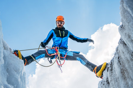 Climber in vibrant gear overcomes over crevasse on icy glacier during ascent under clear sky with snow-covered peaks in background. Extreme active people high-altitude mountaineering conceptの写真素材