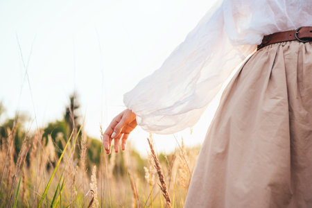 Gently hand of woman in loose blouse and beige skirt walking by golden meadow enjoying sunset warm sunlight peaceful nature atmosphere expressing freedom serenity mindfulness and harmony with natureの写真素材