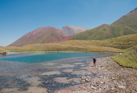 Lonely hiker with a backpack enjoying majestic green hills with unique curved shapes and lakes while trekking in Pamir Range mountains at 3600â¯m altitude. Stunning landscapes of Southern Kyrgyzstan.の写真素材