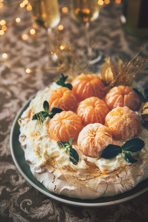 Pavlova cake low light still life pouring with powder sugar meringue-based dessert named after the Russian ballerina decorated with fresh mandarins, caramel, mint, physalis with Christmas lightsの写真素材