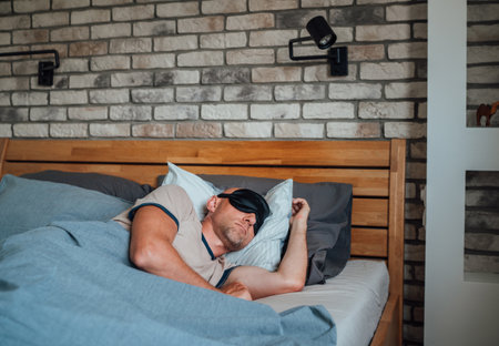 middle-aged bald man Portrait with short beard wearing black sleeping mask lying comfortably in bed during early morning symbolizing healthy sleep, stress relief, relaxation, mentaの写真素材