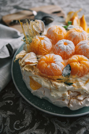 Pavlova cake low light still life pouring with powder sugar meringue-based dessert named after the Russian ballerina decorated with fresh mandarins, caramel, mint, physalis with Christmas lightsの写真素材
