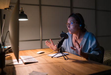 Speaking middle-aged woman in headphones chatting on video call using microphone with pop filter and desktop computer, internet communication technology, online video conference conceptの写真素材