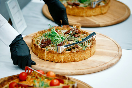Hands of a waiter in black gloves putting pies with sprouts on top, chopped into pieces with metal tongs on the banquet table where there are already other piesの写真素材