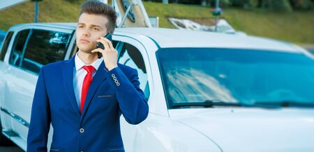 A young attractive business man in a blue business suit with a red tie talking on the phone near an expensive white car with an open doorの写真素材