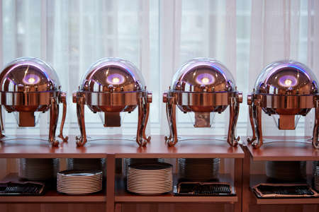 Round food warmers standing on a table above shelves with dishes and trays with forks and knives