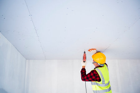 A young Asian construction worker in safety clothing and work gloves is fastening the drywall ceiling to the metal frame using an electric screwdriver on the ceiling covered in shiny aluminum foil.の写真素材