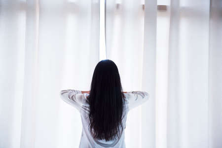 A beautiful Asian woman wakes up in the morning, the sun shines on her from the large window in the bedroom. The young girl welcomed the new day with a warm sunshine.の写真素材