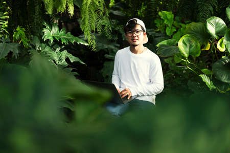 Young Asian man using a laptop computer Happy young Asian man working on his laptop outside the house under a green tree.の写真素材