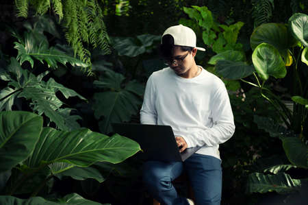 Young Asian man using a laptop computer Happy young Asian man working on his laptop outside the house under a green tree.の写真素材