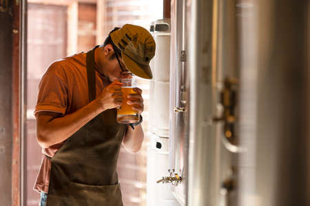 A young man works in a brewery and checks the quality of craft beer. The brewery owner tastes the best beers from Bach. A man's shortcut fills a glass of beer withの写真素材