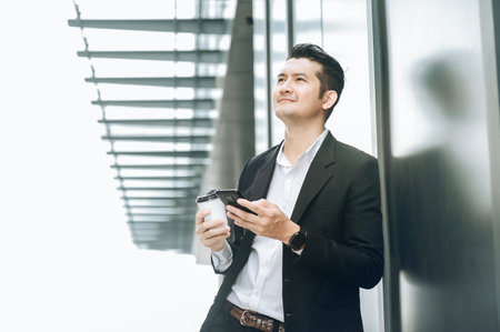 Handsome smiling asian businessman holding smartphone in office Businessman looking at hand holding coffee cup while standing alone in modern workplace.の写真素材