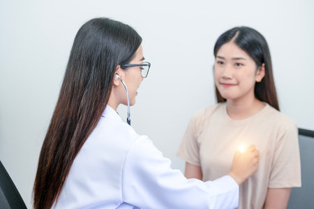 Asian female doctor in uniform holding a stethoscope and using it to measure the heart rate of an asian female patient with heart diseaseの写真素材