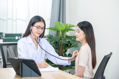 Asian female doctor in uniform holding a stethoscope and using it to measure the heart rate of an asian female patient with heart diseaseの写真素材
