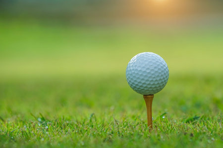 Closeup golf ball on tee ready to be shot. Golf ball on tee in the evening golf course with sunshine. Blurred set of golf clubs over green field background.の写真素材