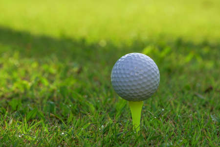 Closeup golf ball on tee ready to be shot. Golf ball on tee in the evening golf course with sunshine. Blurred set of golf clubs over green field background.の写真素材