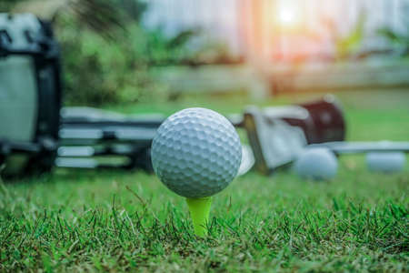 Closeup golf ball on tee ready to be shot. Golf ball on tee in the evening golf course with sunshine. Blurred set of golf clubs over green field background.の写真素材