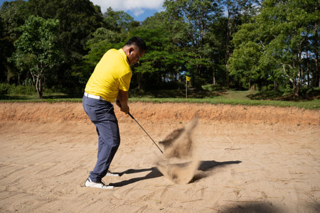 Asian golfer swings in a sand pit during pre-match practice at a golf course. A professional golfer hitting his ball out of a bunker with the sand and ball in mid-air.の写真素材