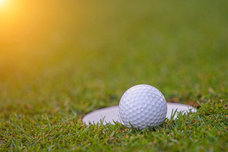 Golf ball on green grass in the evening golf course with sunshine background. Golf ball on the edge of hole on the green grass with warm tone and sunset.の写真素材
