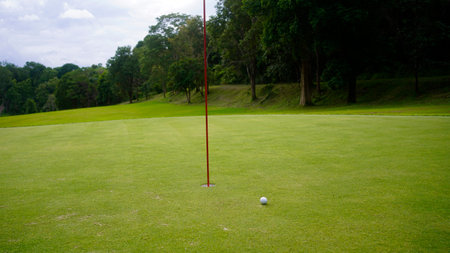 Golf ball is on a green lawn in a beautiful golf course with morning sunshine. golf ball is on the putting green next to the hole with the flagpole.の写真素材