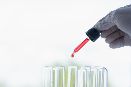 Chemist hand holding a medical dropper filled with red solution dripping chemical drops into a laboratory scientific cylinder full of red liquid for an experiment in a science research labの写真素材