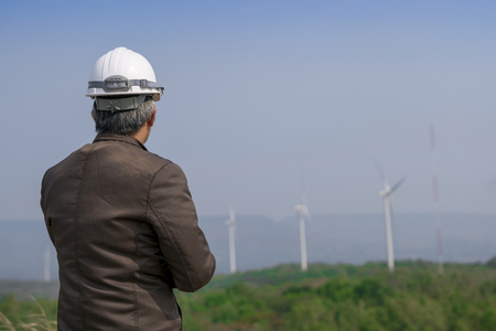Senior engineer man with safety helmet looking to wind turbine for checking its working on the high hill mountain.Wind turbine electric generator,clean energy concept.の写真素材
