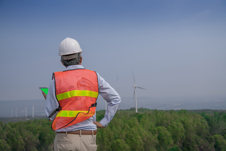 Senior engineer man with safety helmet looking to wind turbine for checking its working on the high hill mountain.Wind turbine electric generator,clean energy concept.の写真素材