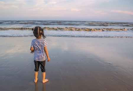 Asian little girl standing on the beach in sunset. Eyes looking to the sea. Image from behind the girl. Beautiful sunset time, reflex on the beach and colorful sky.の写真素材