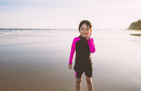 Attractive adorable Asian little girl in long sleeve swimming suit on the beach in sunset. Girl shows victory gesture for confidence. Big smiling face. Space for text.の写真素材