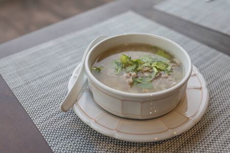 Rice soup in a white bowl on table. Natural light in a morning. Breakfast with rice soup with pork, topping with green vegetable - coriander and celery.の写真素材