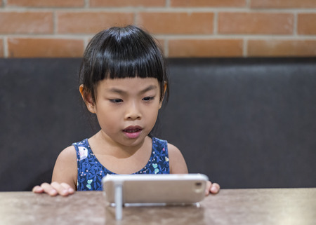 Little child or Little girl watching movie in smart phone. Smart phone with stand on wood table. Sitting on sofa. Brick wall as backdrop.の写真素材