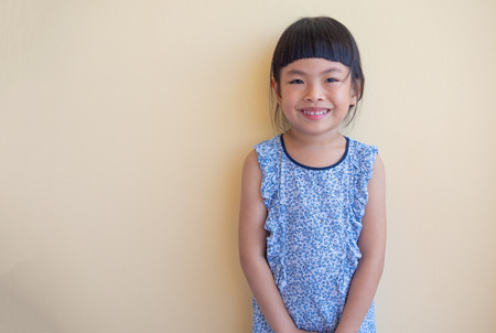 Cute Asian little girl in blue cute dress. Smiling face. Looking at camera. Background earth tone wall.の写真素材