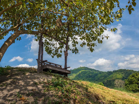 Old wooden swing under a tree on hill. Background of cloudy sky and mountain.の写真素材