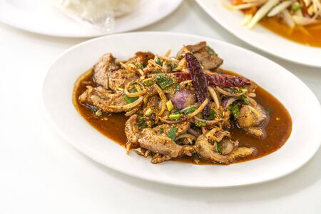 Nam Tok, Spicy BBQ pork or beef salad of traditional Eastern Thailand food. Food is in white plate on white table. Blurred background of Som Tum or papaya spicy salad and stick rice.の写真素材