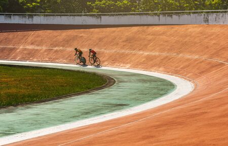 Two Asian bicyclists on cycle track outdoor velodrome. Racing at old orange velodrome in Bangkok, Thailand. Professional cyclists practice together before competition on a hot day under sun.の写真素材