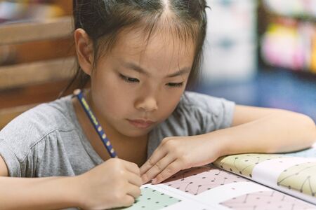 Portrait cute Asian child girl at home, doing homework or lesson, focus, concentrate to lesson, hair tie, holding pencil, lesson book on table. Self education school child girl or homeschool.の写真素材