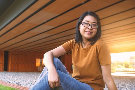 Portrait middle-aged Asian woman with eyeglasses sits on ground under shadow of building inside the park. Wearing casual clothes. Evening's golden light shines through the building behind the woman.の写真素材